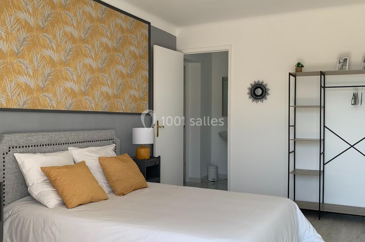 Chambre lumineuse avec lit double, coussins jaunes, tableau décoratif, étagère et porte ouverte sur une salle d'eau.