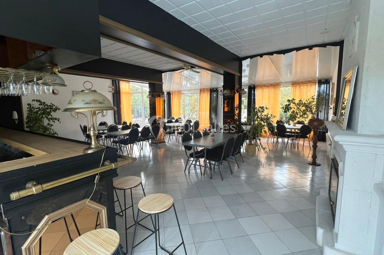Salle de restaurant lumineuse avec tables dressées, chaises noires, bar en bois et grandes baies vitrées avec rideaux jaunes.