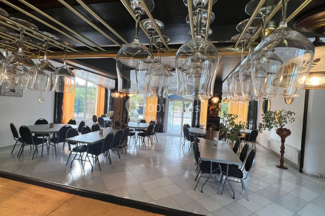 Salle lumineuse avec tables rectangulaires, chaises noires et verrerie suspendue, donnant sur de grandes fenêtres.