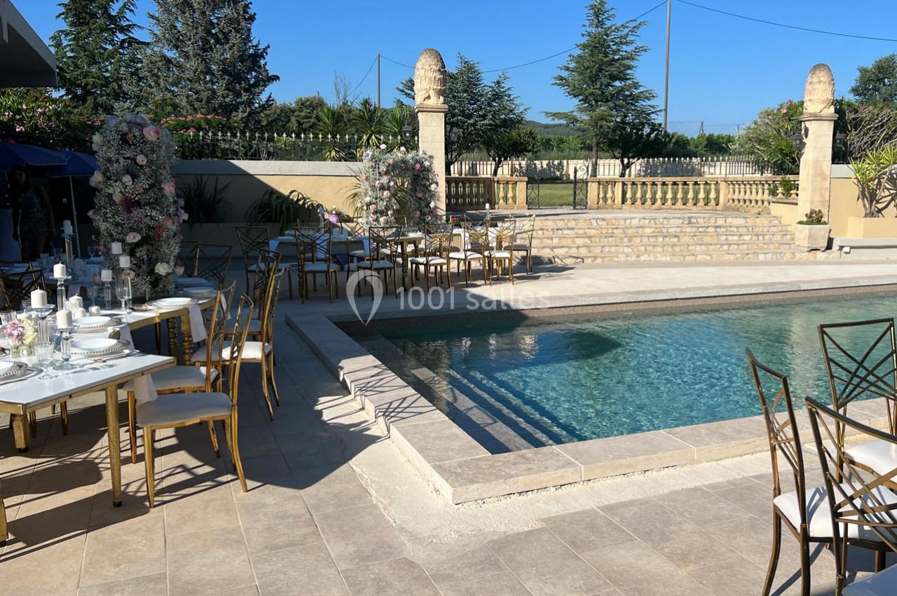 Terrasse avec piscine, tables dressées pour un événement, décor floral et clôture en pierre en arrière-plan.