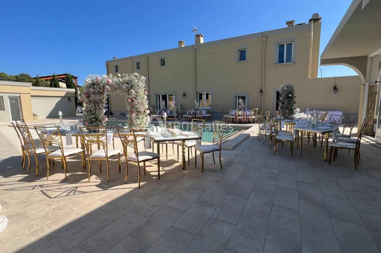 Terrasse aménagée pour un événement avec tables, chaises dorées et arches fleuries près d'une piscine.