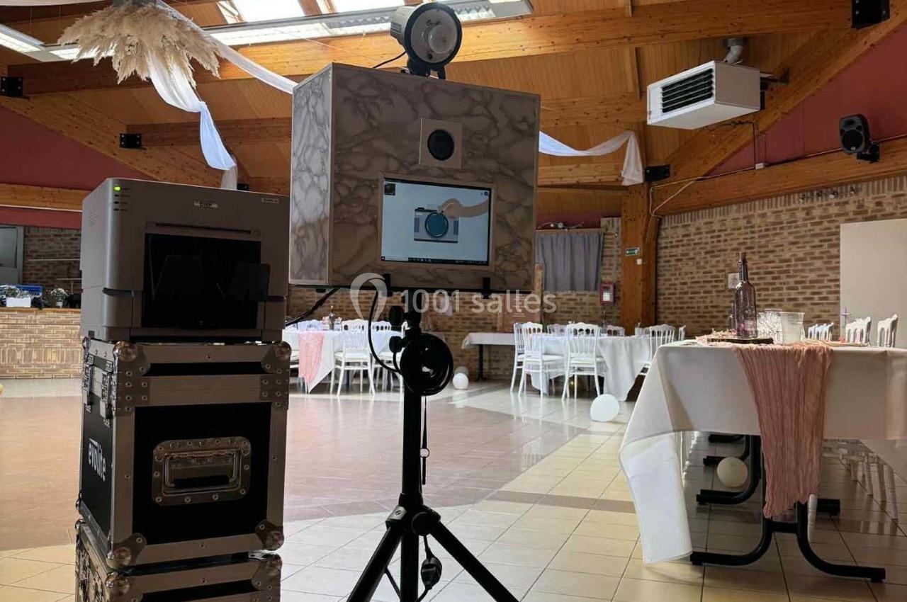 Borne photo installée dans une salle de réception décorée, avec tables dressées et éléments suspendus au plafond.