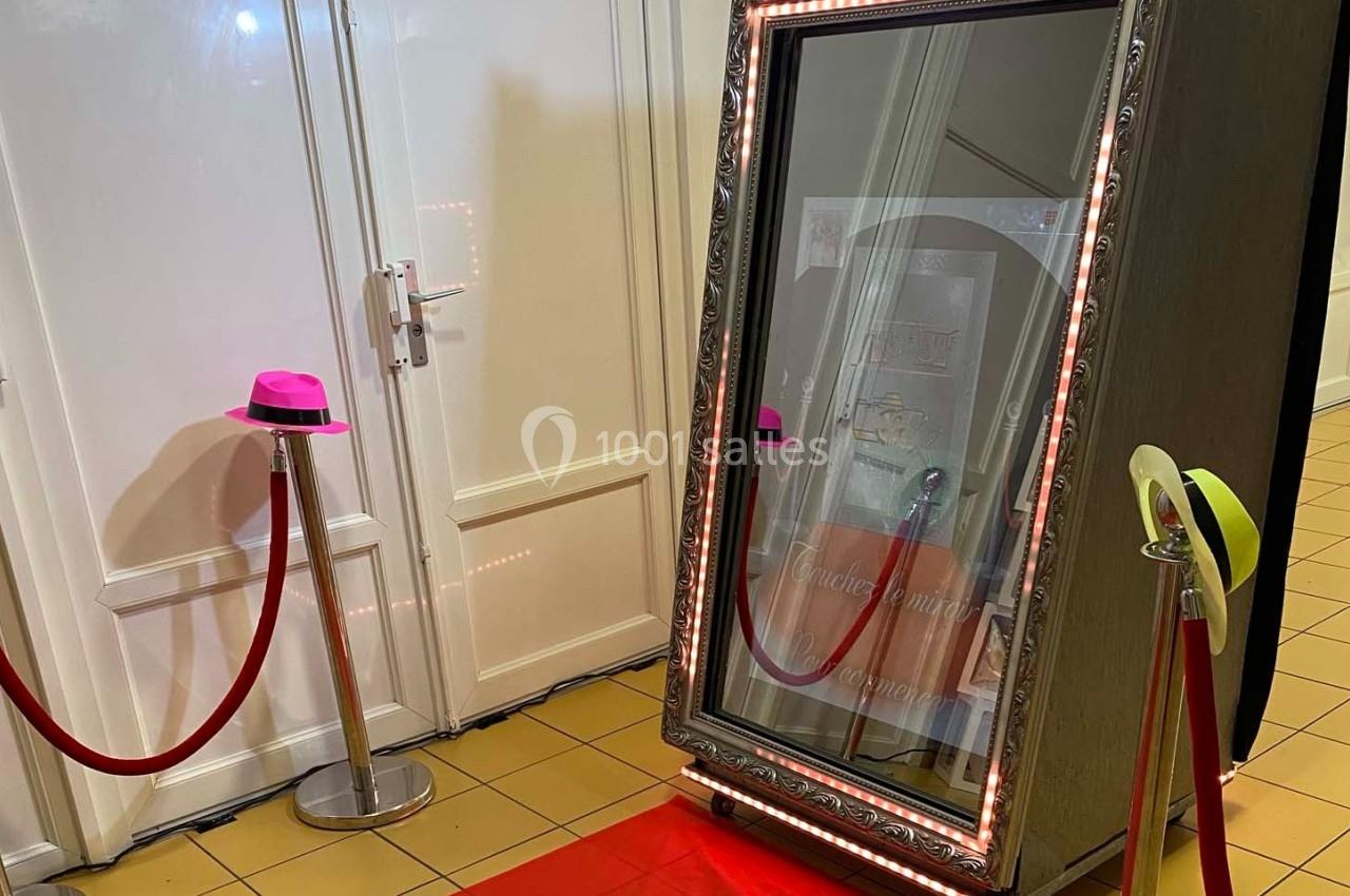 Miroir interactif encadré de lumières, posé sur un tapis rouge avec des chapeaux colorés à proximité.