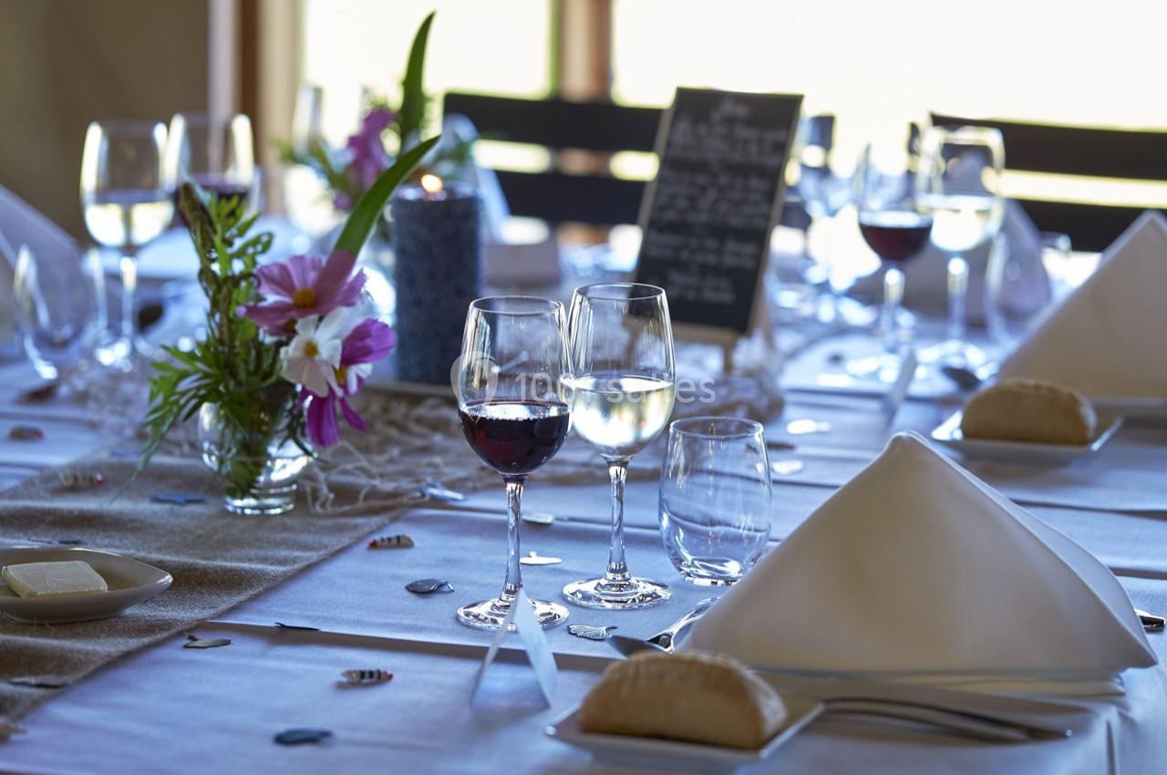 Table dressée avec nappes blanches, verres de vin, fleurs, serviettes pliées et menu sur ardoise en arrière-plan.