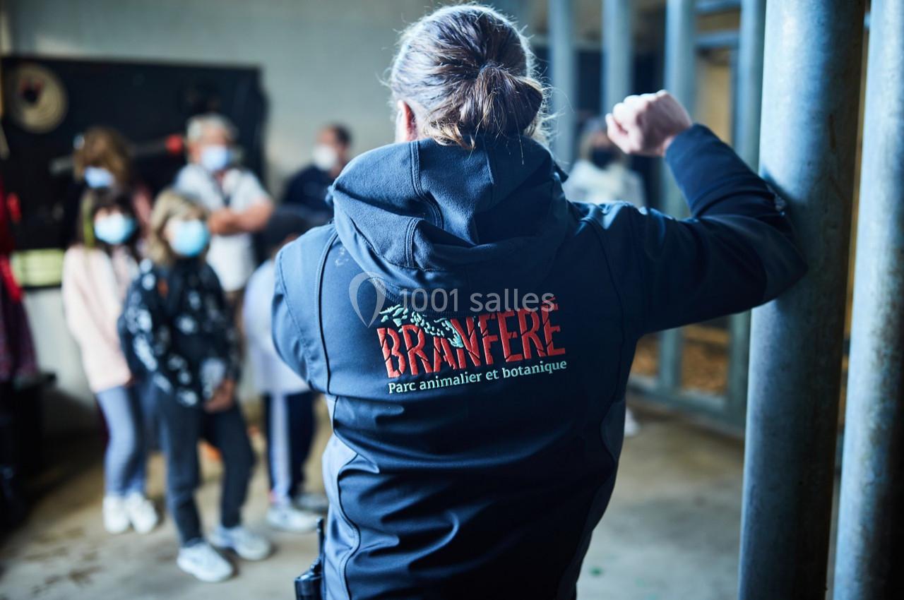 Une personne de dos portant une veste ’Branféré’ face à un groupe de visiteurs dans un espace intérieur.