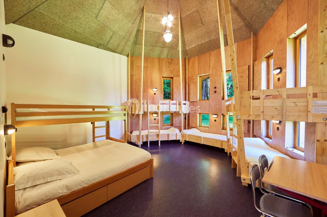 Dortoir en bois avec lits superposés, grandes fenêtres, éclairage suspendu et mobilier simple.