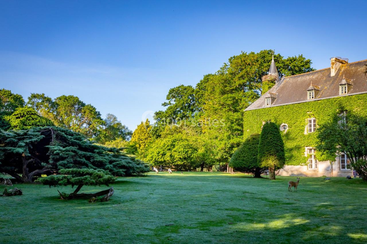 Manoir couvert de lierre entouré d'un grand jardin verdoyant avec des arbres et un chevreuil au loin.