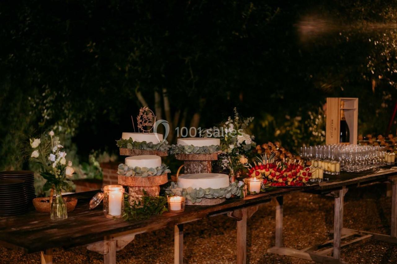 Table en bois décorée pour un événement, avec gâteaux, bougies, fleurs et verres disposés sous un éclairage tamisé.