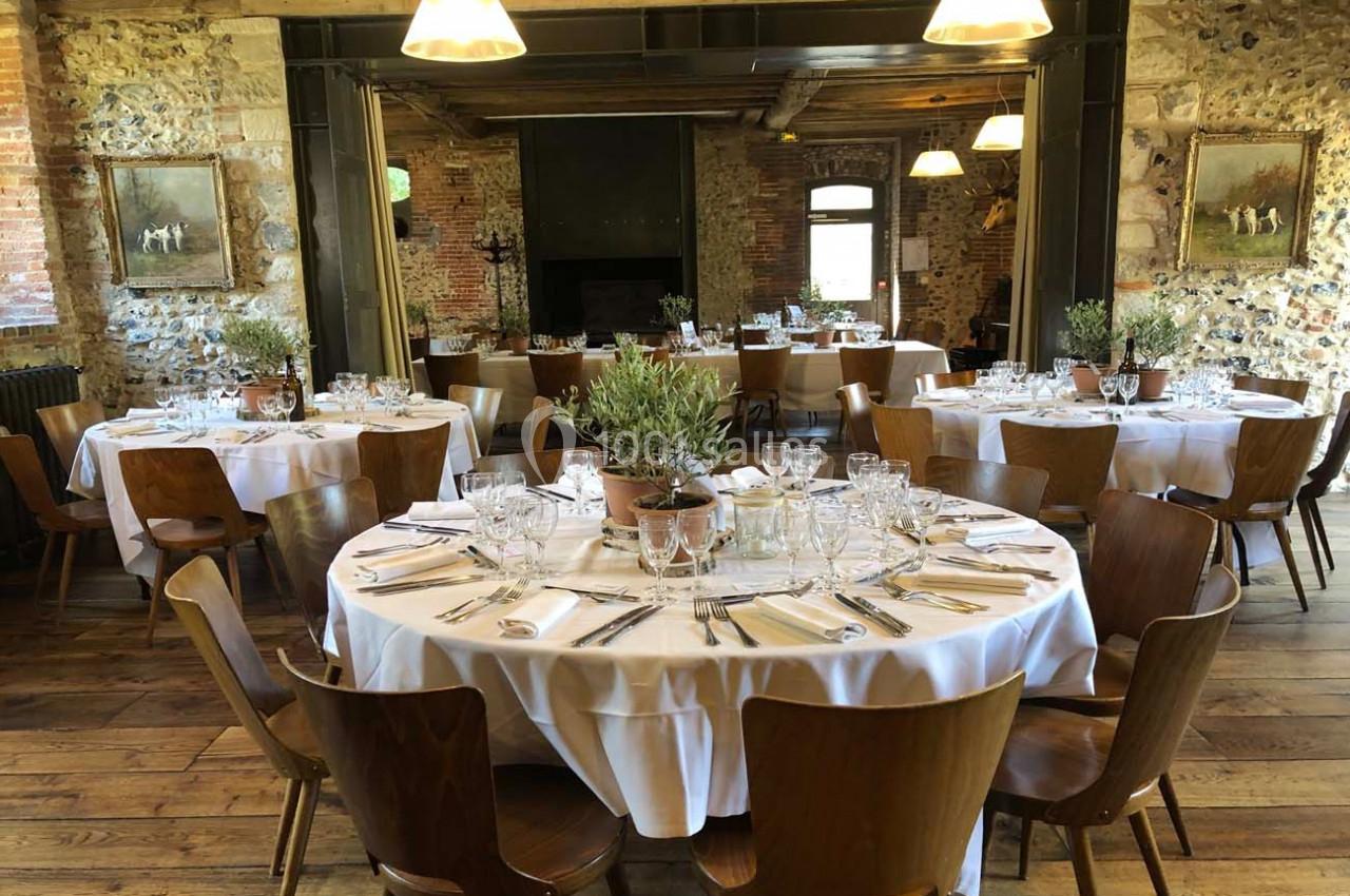 Salle de réception avec tables rondes dressées, nappes blanches, chaises en bois et décorations végétales.