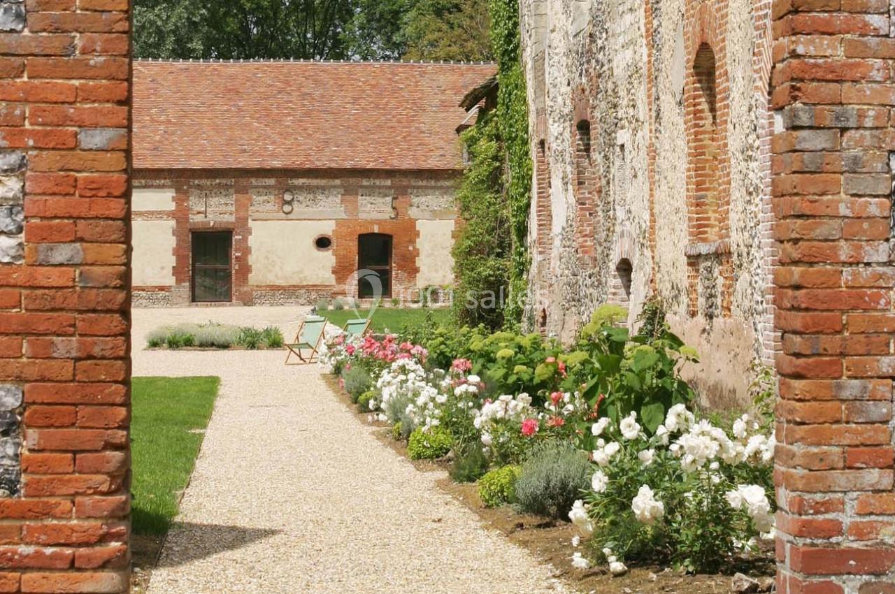 Cour intérieure avec allée gravillonnée, parterres fleuris et bâtiments en briques et pierres sous un toit en tuiles.