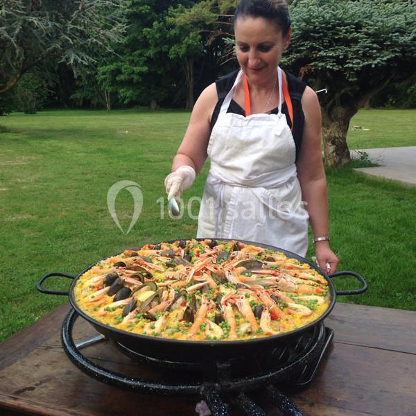Une femme en tablier prépare une grande paella aux fruits de mer sur un réchaud en plein air, dans un jardin verdoyant.