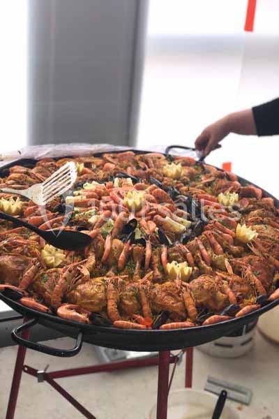 Grande poêle de paella garnie de crevettes, moules, poulet et légumes, avec une main tenant une spatule.