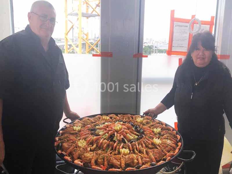 Un homme et une femme présentent une grande paella garnie de fruits de mer dans un cadre lumineux.