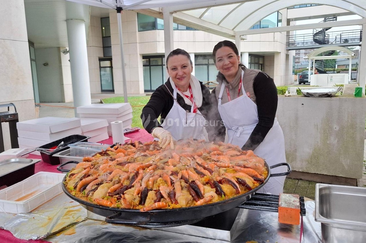 Deux femmes en tablier blanc cuisinent une grande paella fumante sous un chapiteau en extérieur.