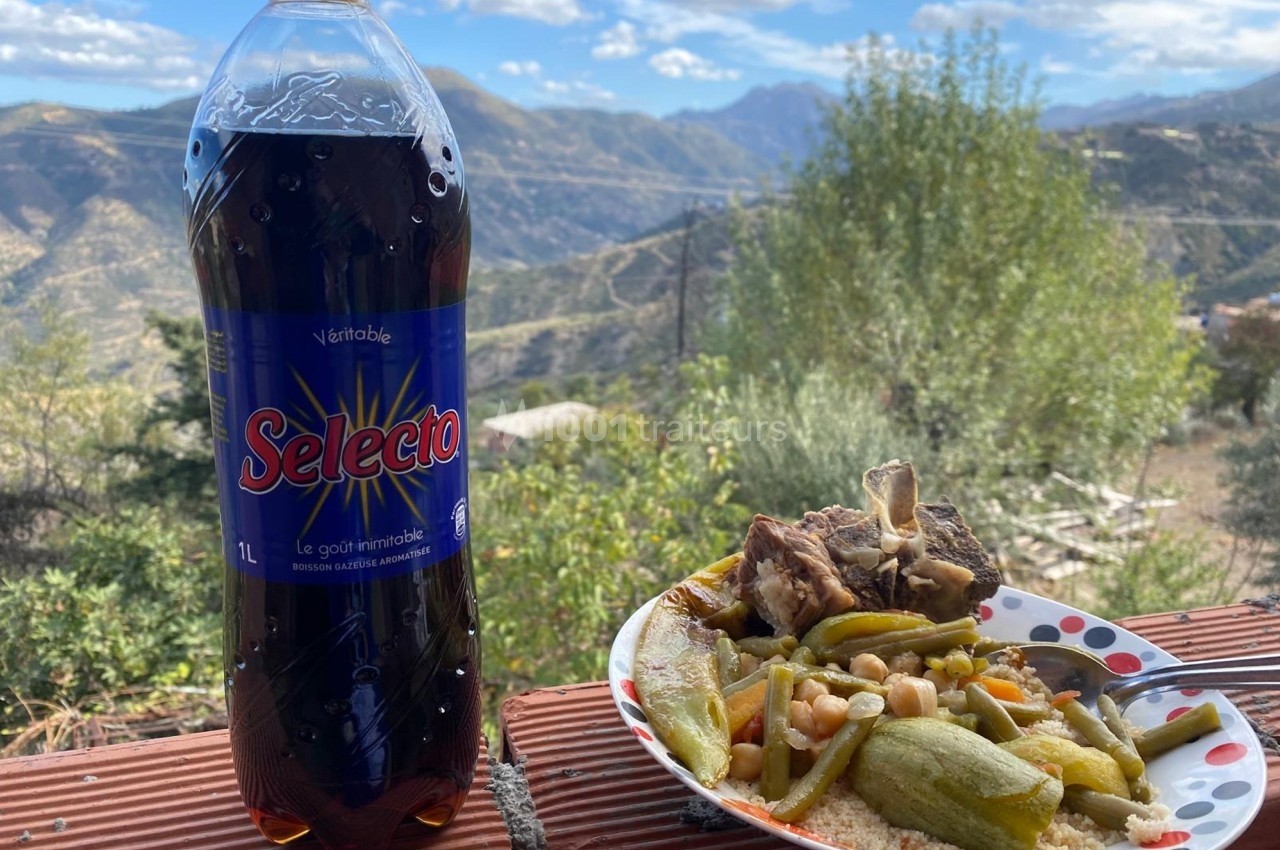 Assiette de couscous avec légumes et viande, accompagnée d'une bouteille de soda, en extérieur avec vue sur des montagnes.