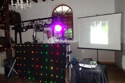 Un DJ mixe derrière une table éclairée de lumières LED violettes, avec un mur décoratif en arrière-plan.