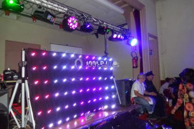 Un DJ mixe derrière une table éclairée de lumières LED violettes, avec un mur décoratif en arrière-plan.