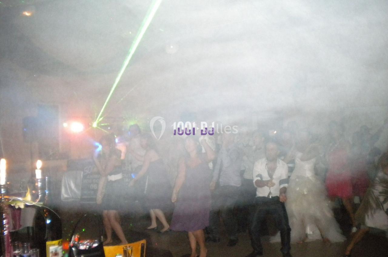 Des personnes dansent dans une salle enfumée avec des jeux de lumière et un DJ en arrière-plan.