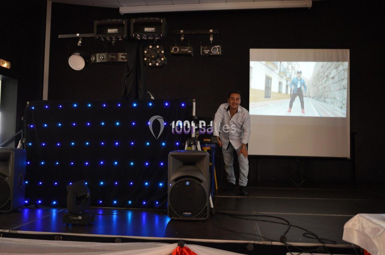 Un homme souriant se tient près d'une table de mixage éclairée, avec un écran projetant une vidéo en arrière-plan.