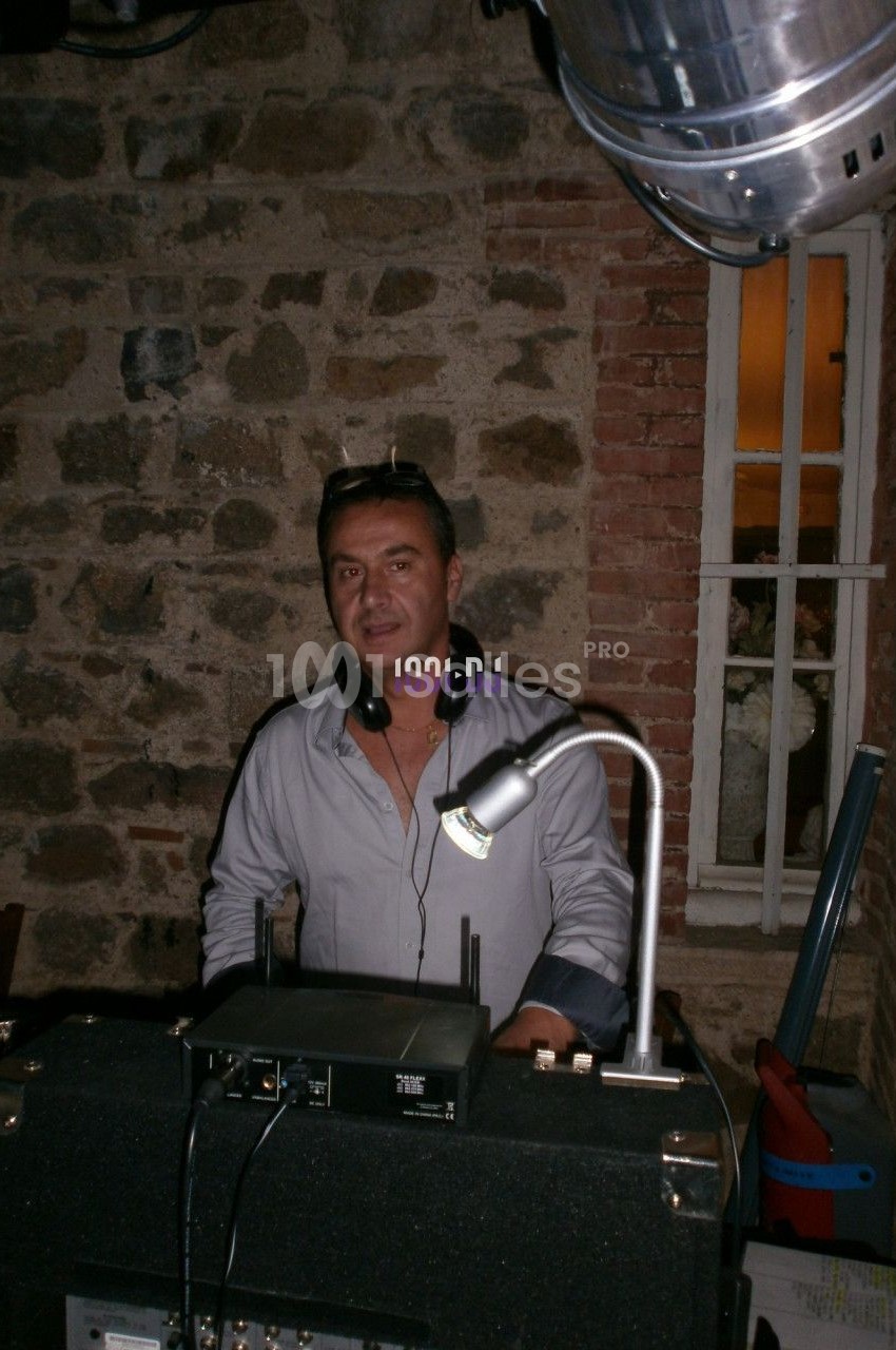 Homme debout derrière une table de mixage dans une pièce aux murs en pierre, éclairée par une lampe.