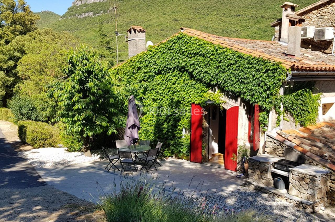 Maison en pierre avec façade recouverte de lierre, volets rouges et terrasse aménagée, entourée de verdure.