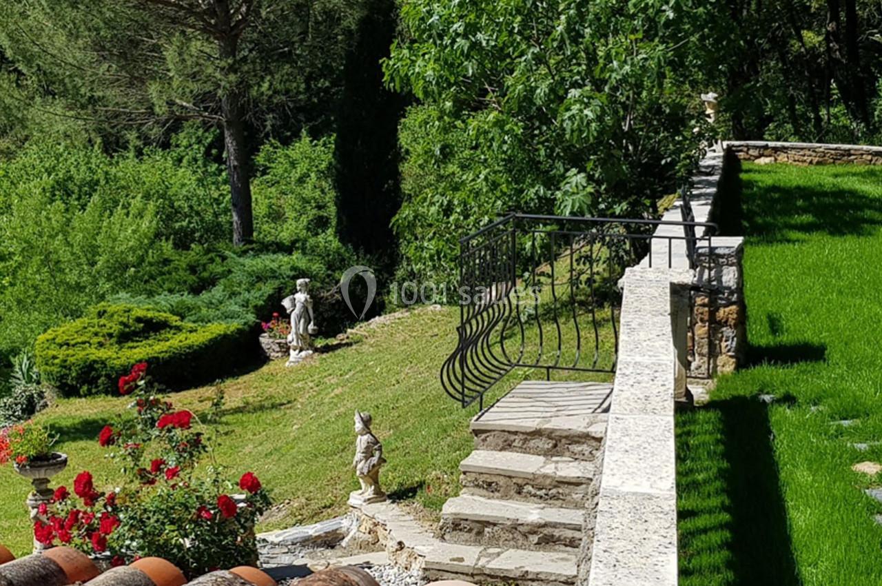 Jardin verdoyant avec statues, escalier en pierre et rambarde en fer forgé, entouré d'arbres et de fleurs rouges.