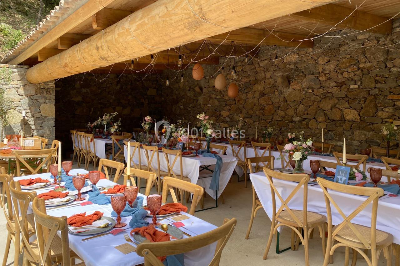 Tables décorées pour un repas festif sous une pergola en bois, avec nappes blanches, serviettes colorées et fleurs.