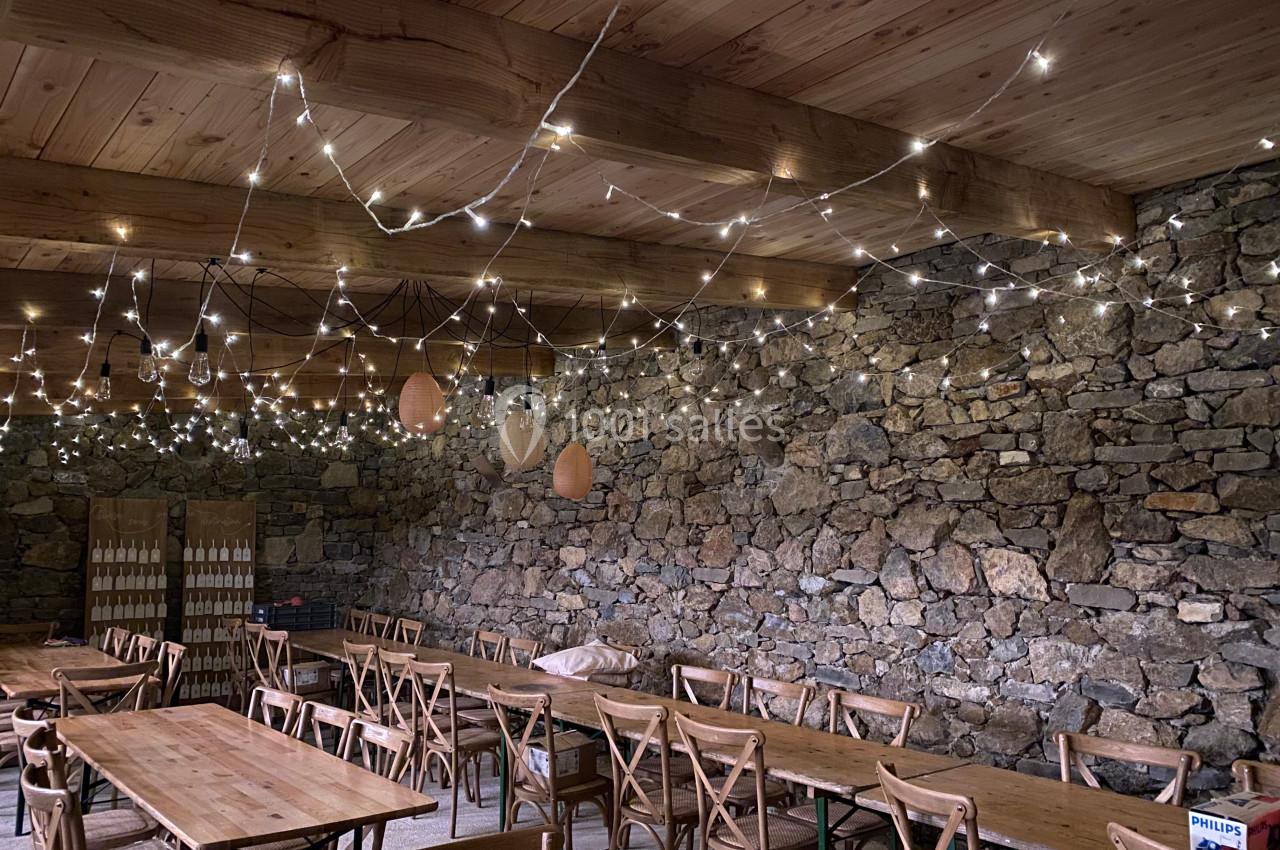 Salle en pierre avec des tables en bois et des chaises, éclairée par des guirlandes lumineuses suspendues au plafond.