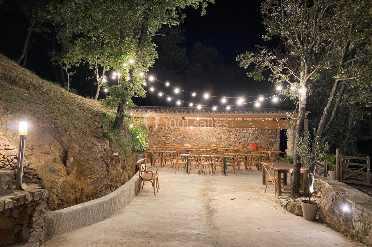 Terrasse extérieure éclairée par des guirlandes lumineuses, avec tables et chaises en bois, entourée de végétation.