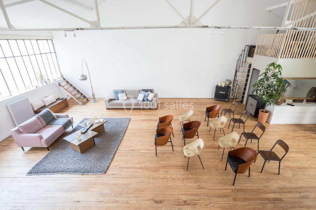 Salle lumineuse avec parquet, canapés, chaises disposées en cercle, grande fenêtre et mezzanine en arrière-plan.