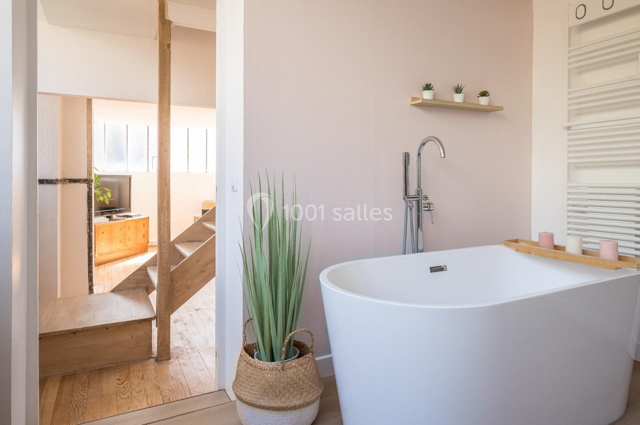 Salle de bain lumineuse avec baignoire blanche, mur rose, plante en pot et vue sur un escalier en bois.