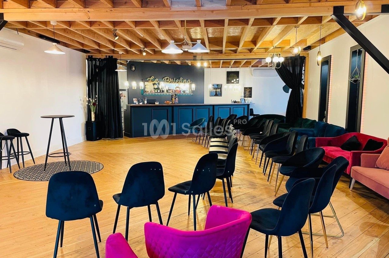 Salle lumineuse avec parquet, chaises disposées en rangées, bar au fond et plafond en bois apparent.