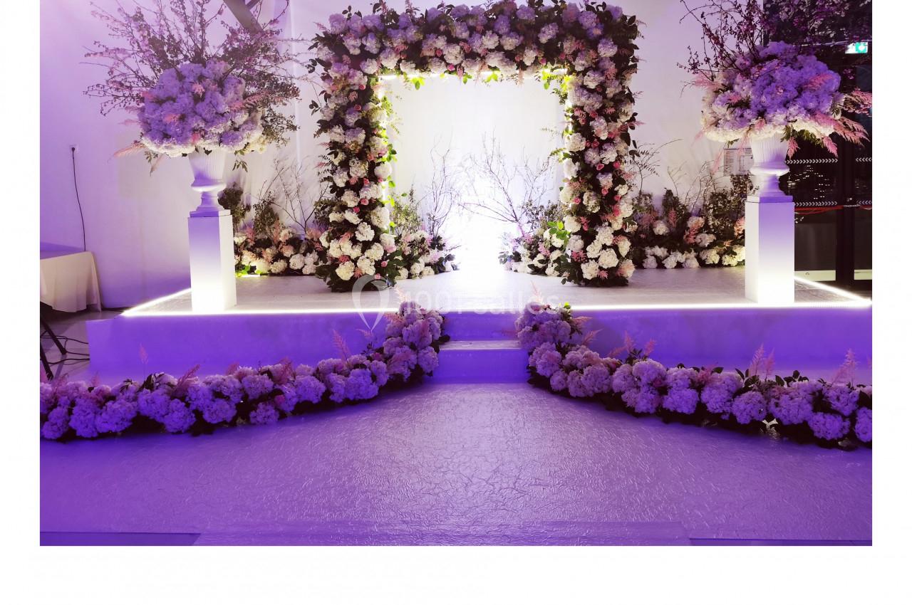 Arche de mariage décorée de fleurs blanches et roses, entourée de compositions florales sur un podium éclairé.