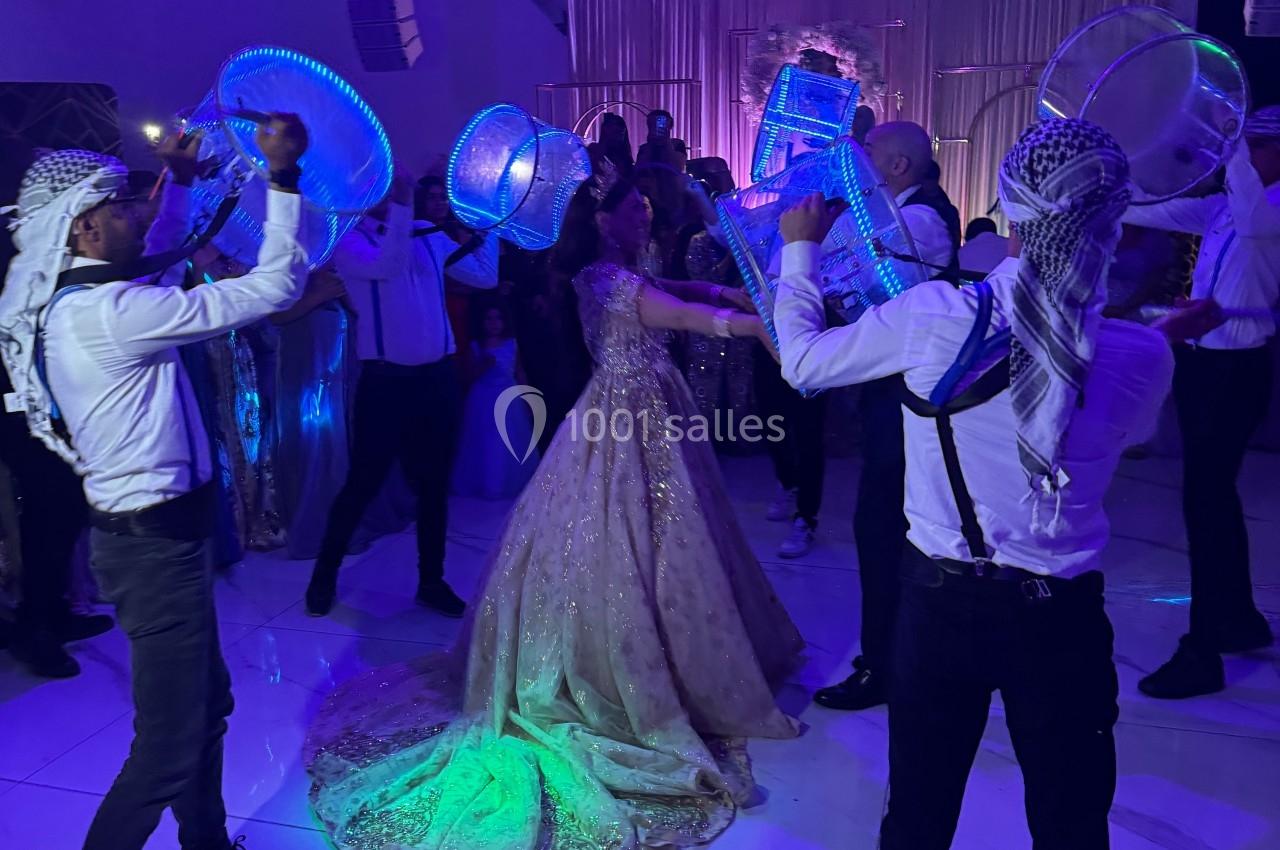 Des danseurs en tenue traditionnelle entourent une mariée sous des lumières colorées dans une salle de réception.