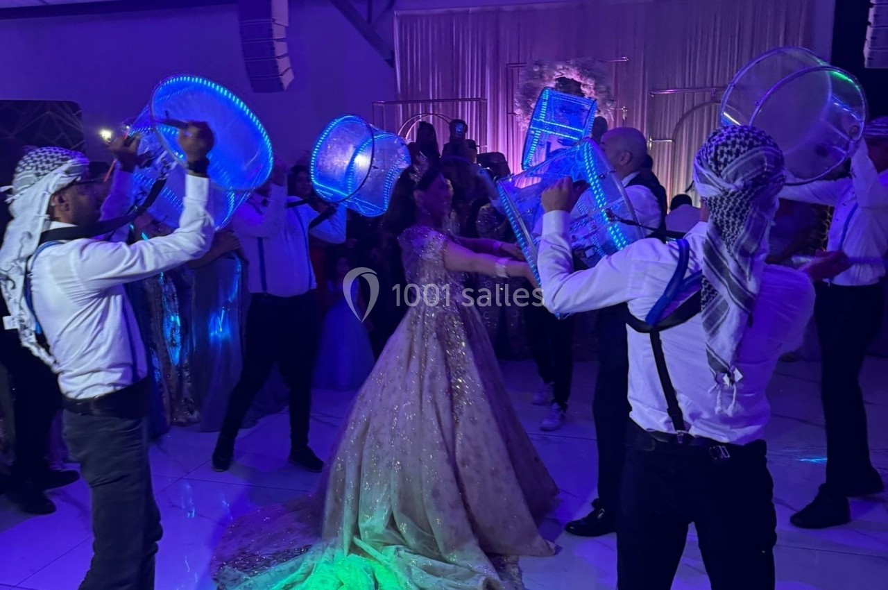 Des danseurs en tenue traditionnelle jouent du tambour lumineux autour d'une femme en robe de soirée dans une salle éclairée.