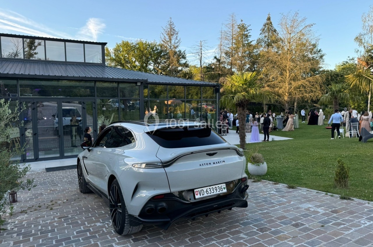 Voiture grise garée sur une allée pavée devant un bâtiment vitré, avec des invités rassemblés dans un jardin.