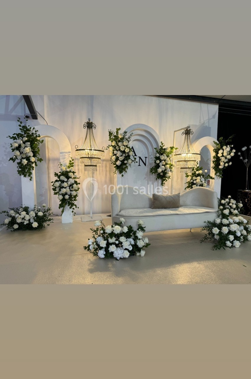 Canapé blanc entouré de compositions florales blanches et vertes, avec chandeliers et décor mural en arrière-plan.