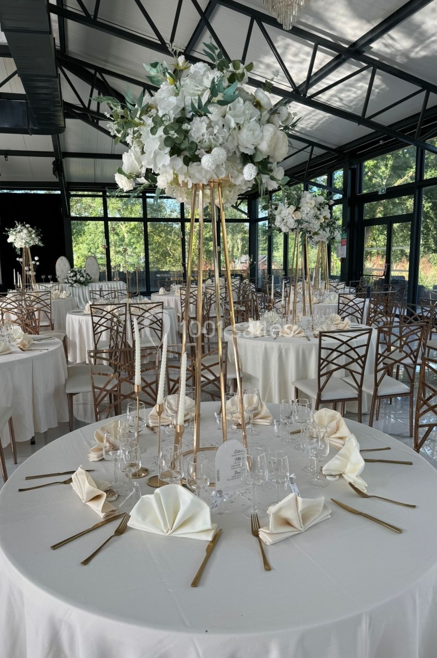 Salle de réception lumineuse avec tables décorées de nappes blanches, centres de table fleuris et couverts élégants.
