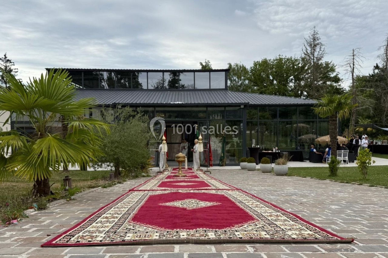 Allée pavée bordée de palmiers menant à une verrière, décorée de tapis rouges et entourée de verdure.