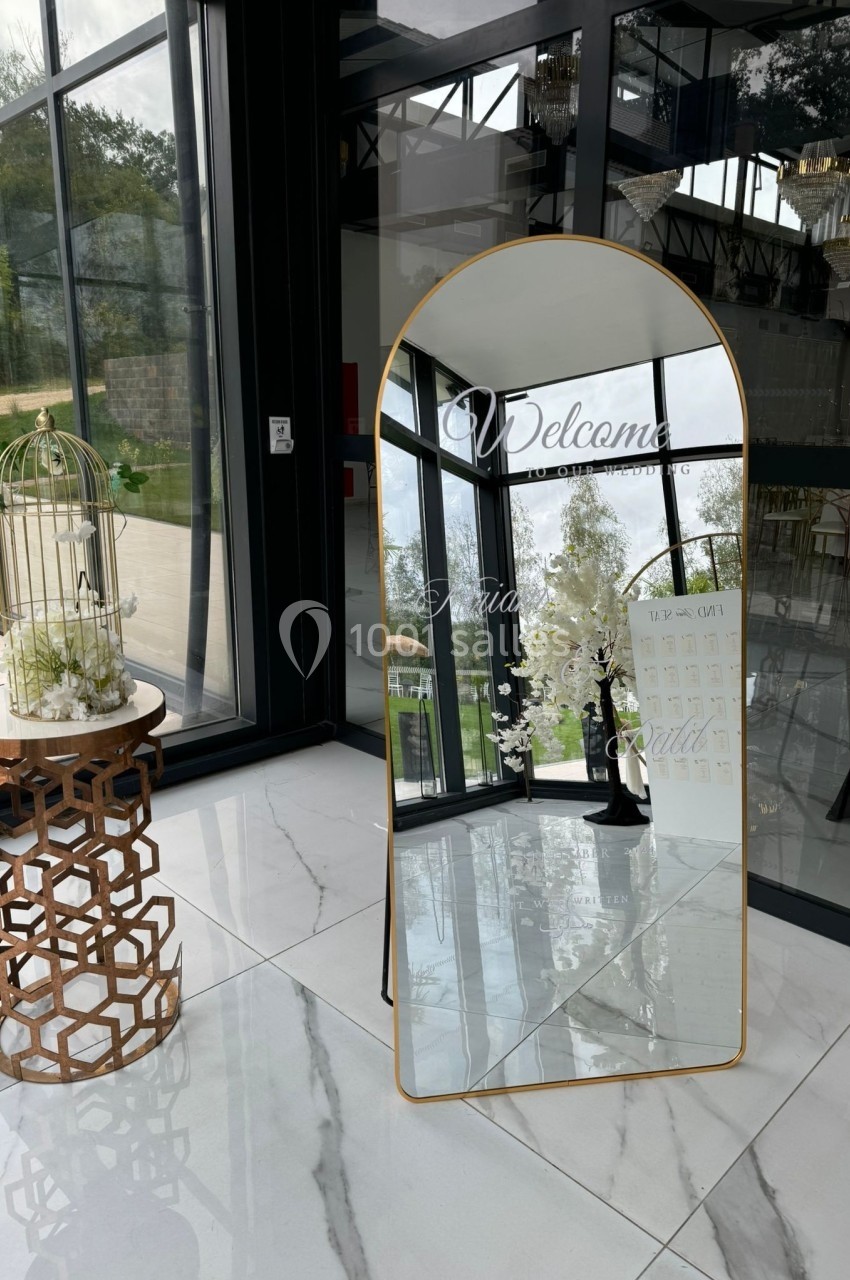 Grand miroir doré avec inscription de bienvenue, placé dans un espace lumineux près d'une table décorative et d'une cage.
