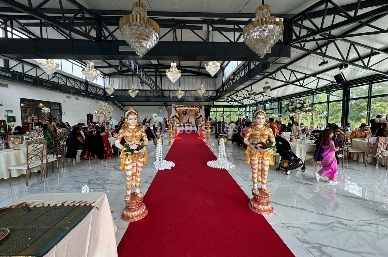 Salle de réception décorée pour un mariage avec tapis rouge, statues traditionnelles et invités assis à des tables.