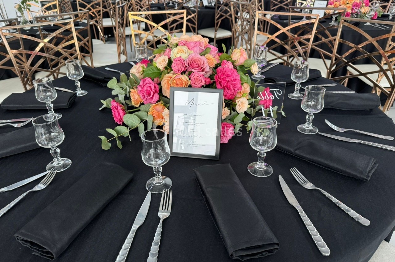 Table dressée avec une nappe noire, couverts argentés, verres, serviettes noires et un centre de table floral coloré.