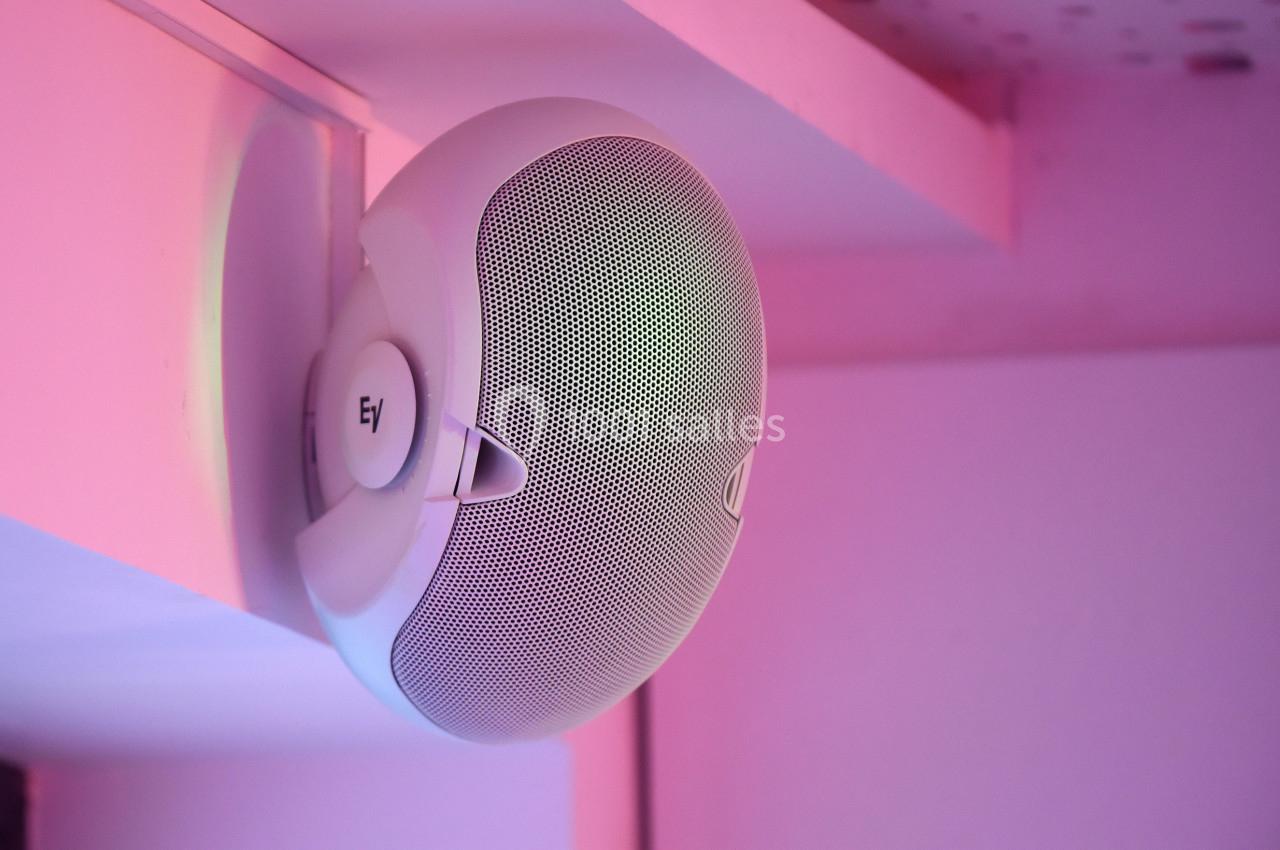 Haut-parleur mural blanc fixé sous une étagère, éclairé par une lumière rose.
