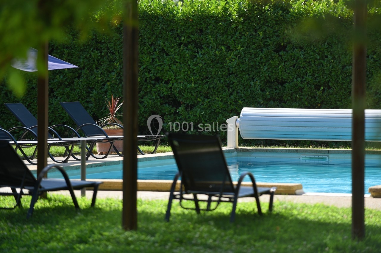 Chaises longues noires autour d'une piscine extérieure entourée de haies vertes, avec un volet roulant partiellement ouvert.