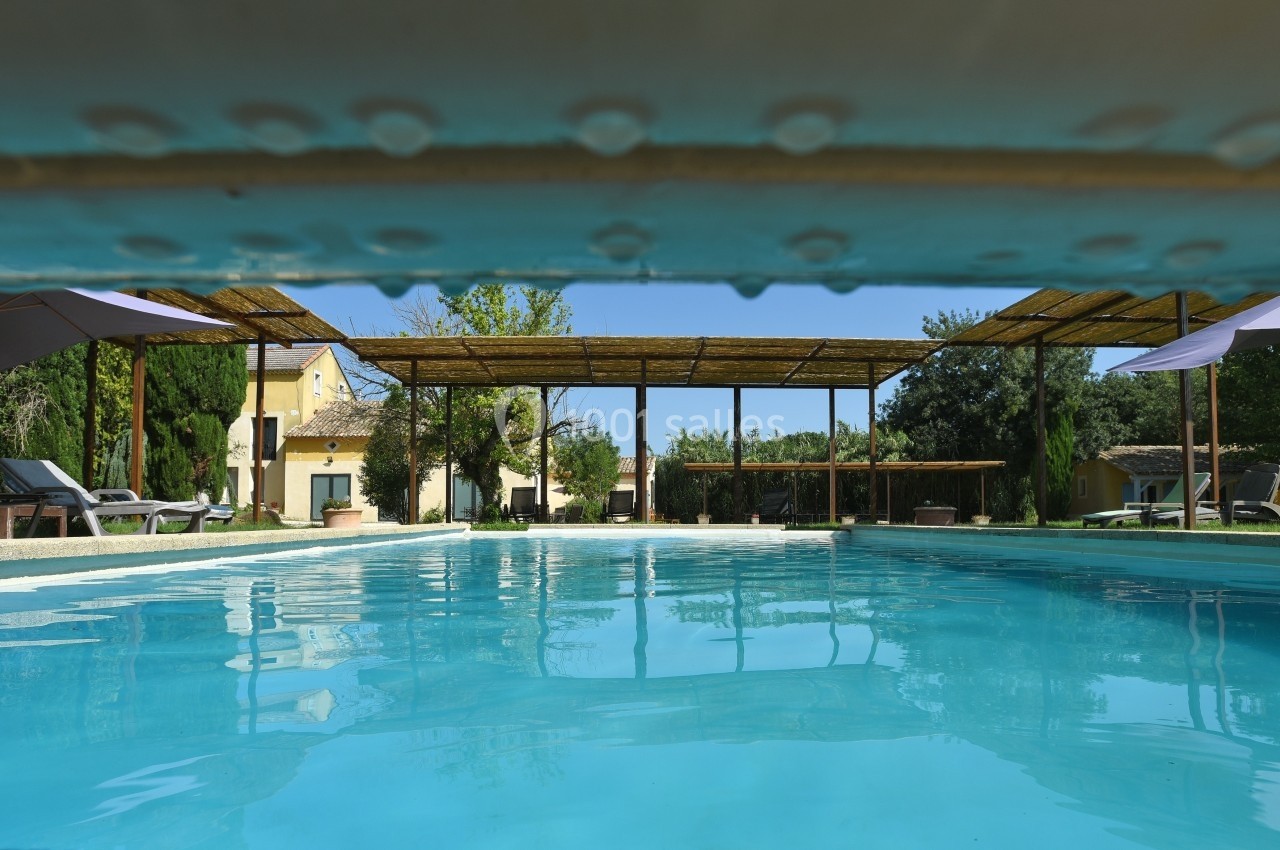 Vue d'une piscine extérieure entourée de transats, parasols et pergolas, avec des arbres et bâtiments en arrière-plan.