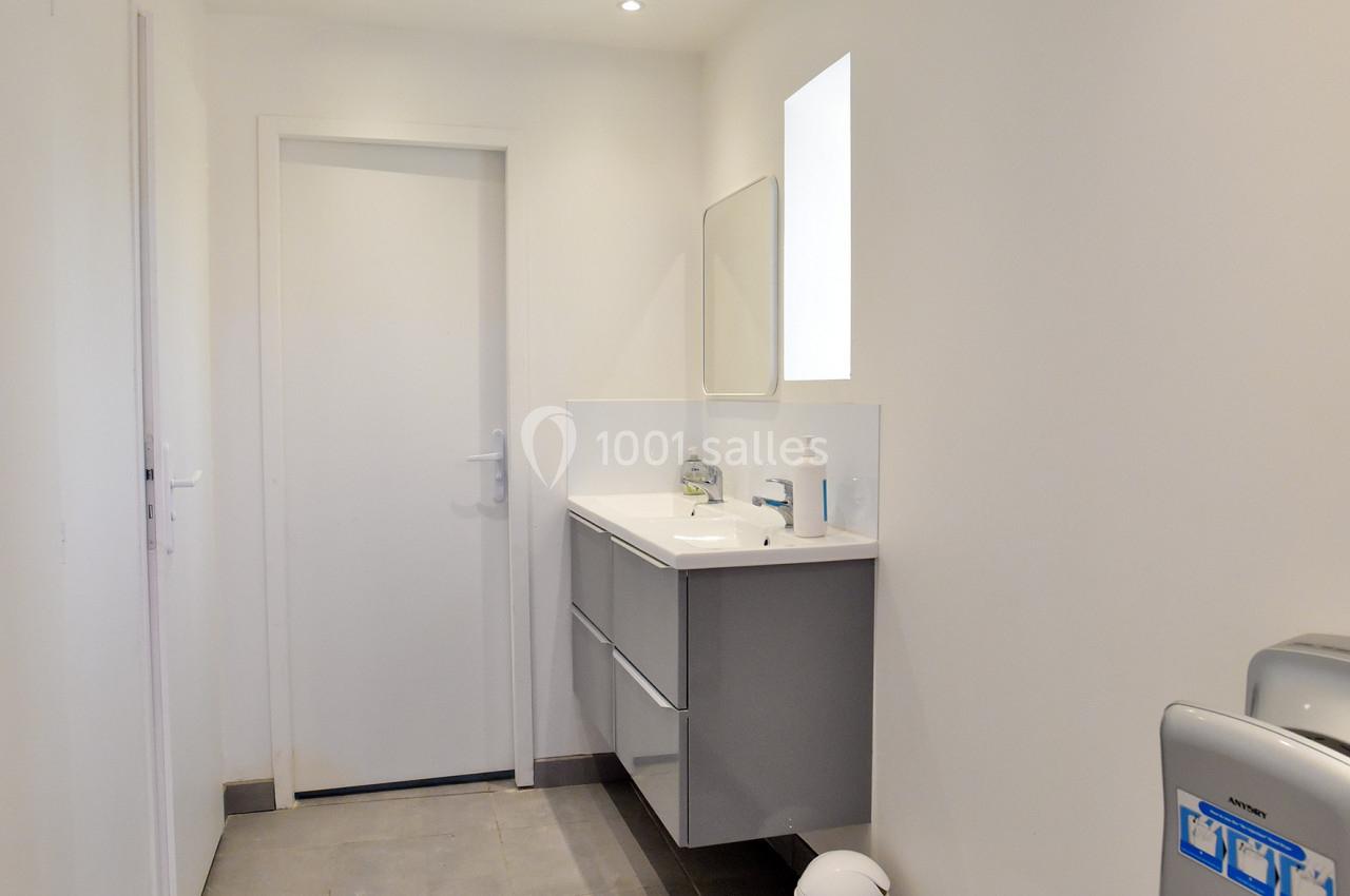 Salle de bain moderne avec carrelage gris, meuble suspendu, miroir, petite fenêtre et poubelle blanche au sol.