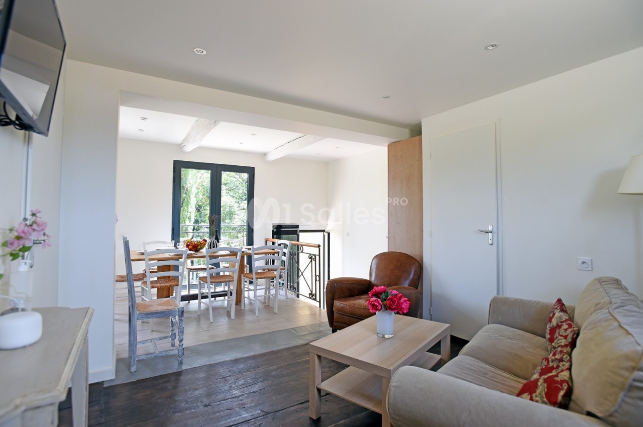 Salon lumineux avec canapé, fauteuil, table basse et vue sur une salle à manger avec table et chaises près d'une fenêtre.