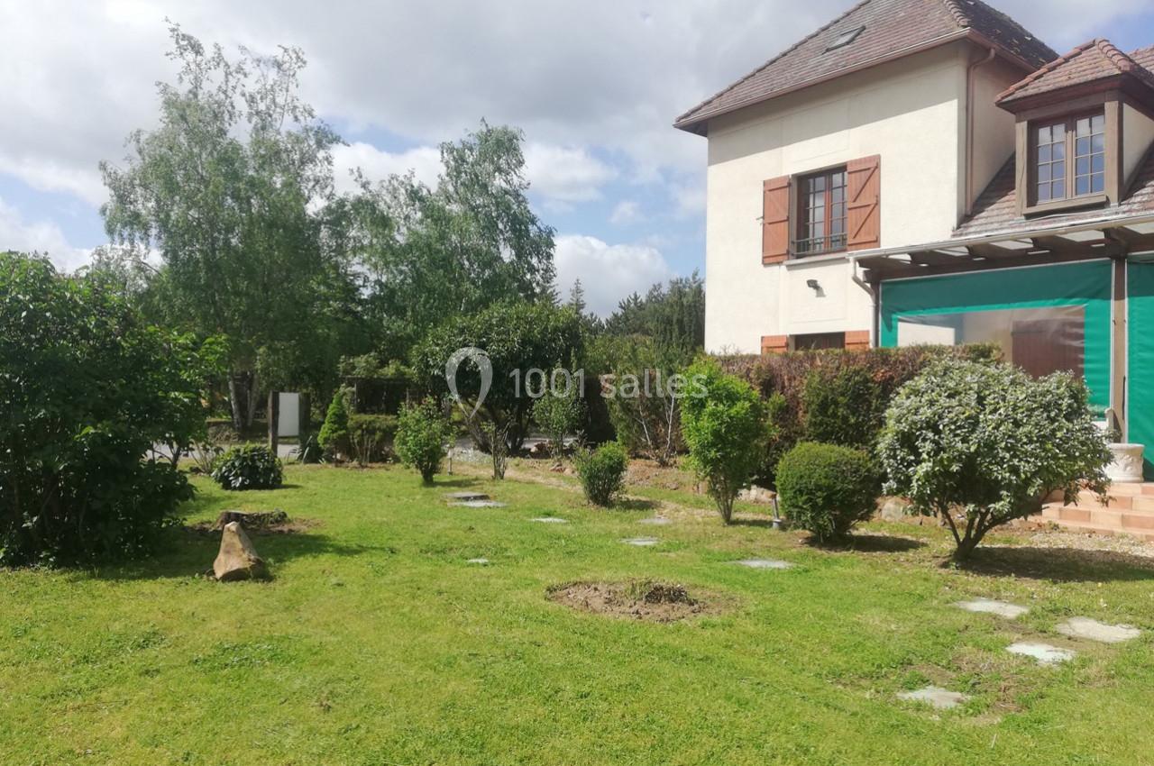 Jardin verdoyant avec pelouse, arbustes et arbres, jouxtant une maison à deux étages avec volets en bois.
