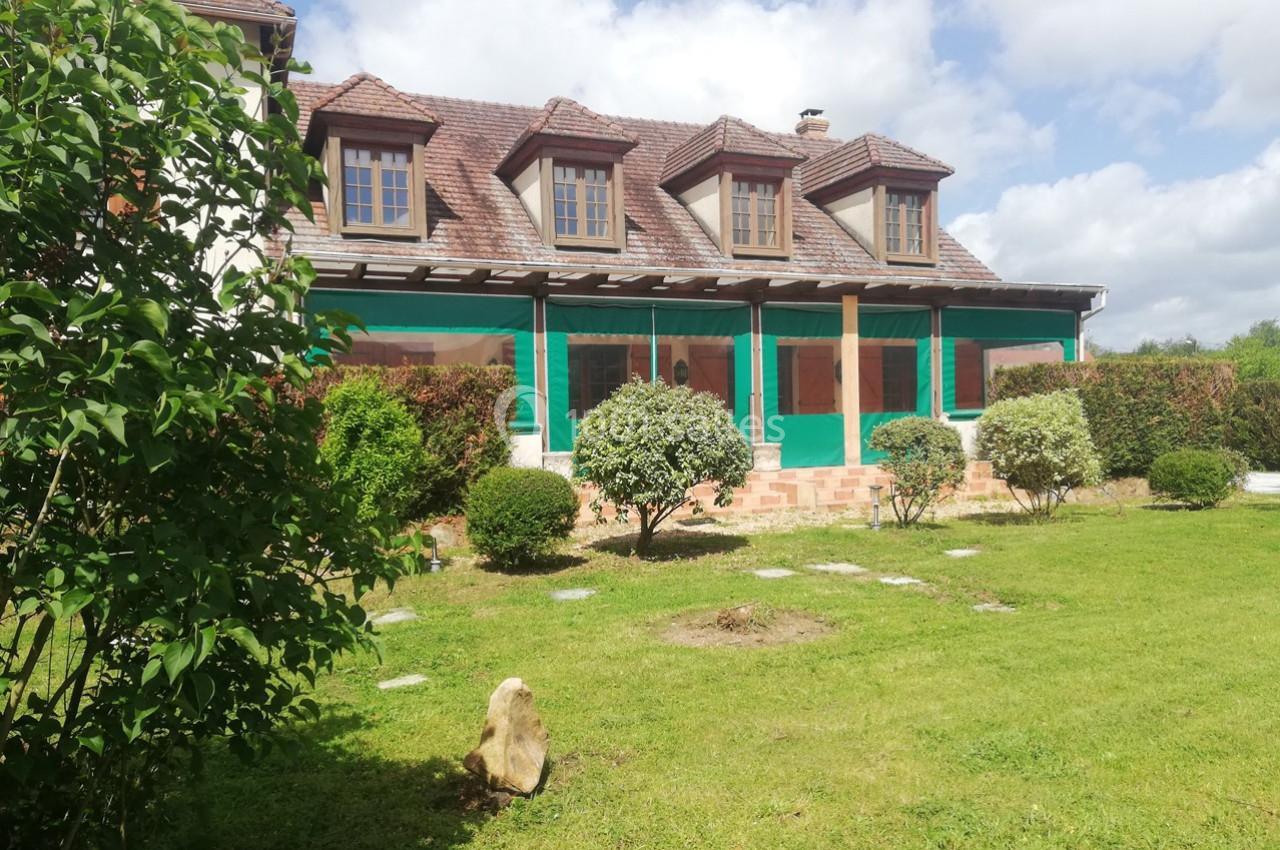 Maison en pierre avec toit en tuiles, entourée d'un jardin verdoyant et d'arbustes sous un ciel partiellement nuageux.