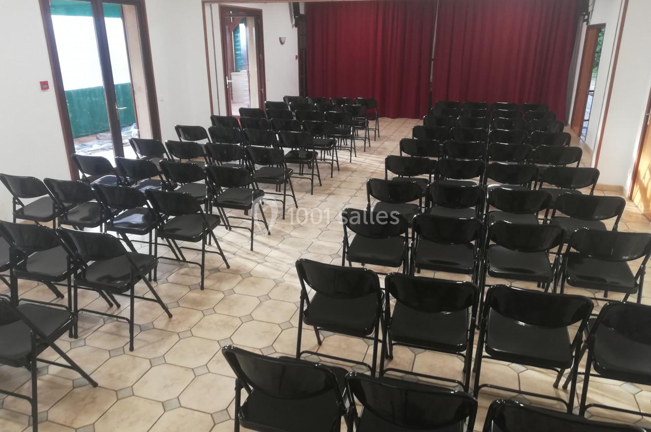 Salle avec des rangées de chaises noires pliantes alignées face à une scène avec rideaux rouges.